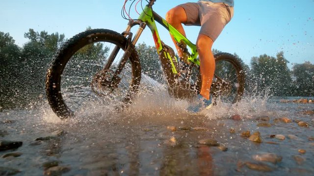 SLOW MOTION TIME REMAP: Unrecognizable male mountain biker splashes the pure river water as he speeds downhill and through tranquil nature on a sunny summer day. Cinematic shot of water drops flying.