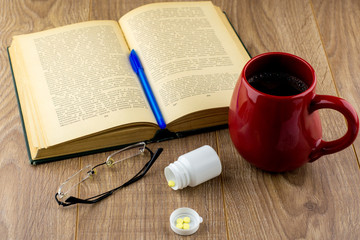 Sleepiness and insomnia: caffeine pills near the pill box, old book, glasses and red large mug of strong coffee on the wooden Desk, blue pen on the book