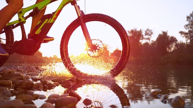 SLOW MOTION, LENS FLARE, CLOSE UP: Unrecognizable cross country cyclist splashes the refreshing river water as he speeds through the picturesque serene nature at sunset. Sportsman riding his bike.