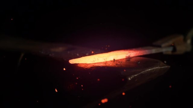 SLOW MOTION, MACRO, DOF: Small black particles flying away from hot red blade while getting forged by an unrecognizable blacksmith. Glowing piece of iron being forged into blade. Forging knife blades.