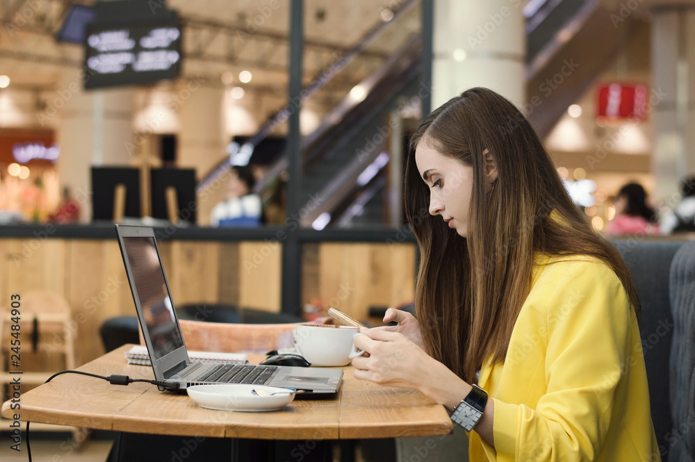 Wall mural young female freelancer in yellow jacket working in a cafe. woman drinking coffee, browsing via mobl