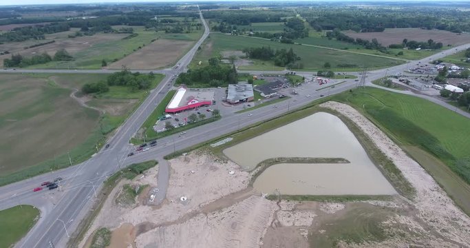 Flying Over Dirty Pond Towards Small Mini Plaza By Street Intersection And Farmers Fields