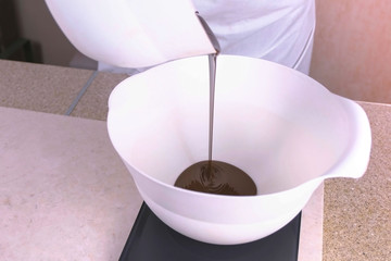 Cook pours liquid dark chocolate in a white bowl, standing on the scales. Close-up view.