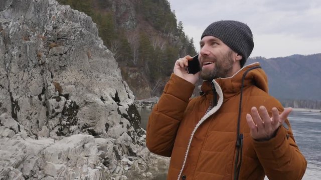 bearded tourist is having video conference with friends on a mobile phone on the bank of a mountain river. slow motion