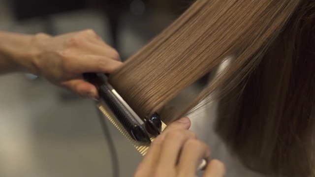 Hairstyling long hair with hair iron in professional beauty studio. Hairstylist doing straightening for long haired woman in barbershop. Hairdresser creating hairstyle brunette woman.