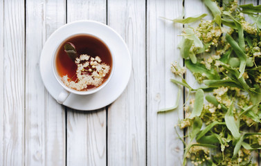 White cup and dish of linden tea on linden blossom background. Herbal medicine concept. Top view. Rustic style.