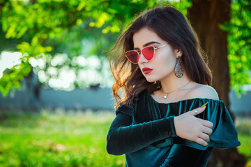 Portrait of beautiful young brunette woman with bright makeup, wearing emerald green velvet dress and bright red sunglasses, standing outdoors with green trees on background.