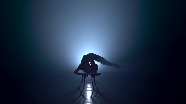 Gymnast on the table sits on the splits. Smoke background. Slow motion. Silhouette