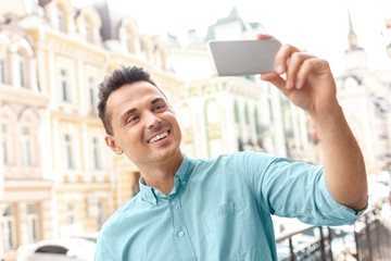 Capturing moments. Young man standing on city street taking selfie on smartphone looking camera cheerful close-up