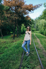 Young hipster girl in blue jeans and fashionable top is standing on railway tracks in the park. Summer photoshoot  outdoors for lifestyle concept. Blonde woman in cool glasses.