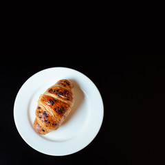 Delicious dessert-crispy croissant with chocolate drops on a white plate stands on a black background. Top view