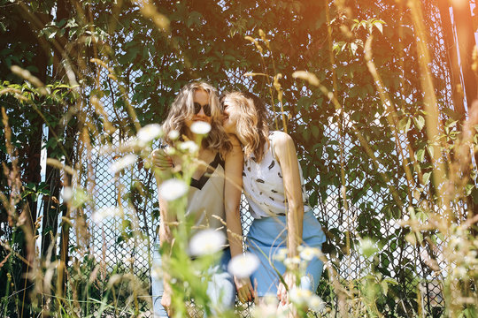 Two hipster girls are standing near the ivy leaves fence outdoors. Funny happy emotions of sisters. Best friends photo session.Stylish and fashionable women. Travel lifestyle concept.