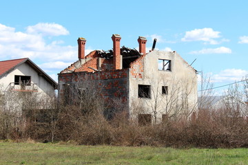 Fototapeta na wymiar Family house bombarded during war with missing windows and completely destroyed roof with only chimneys left untouched surrounded with overgrown underbrush and cloudy blue sky background