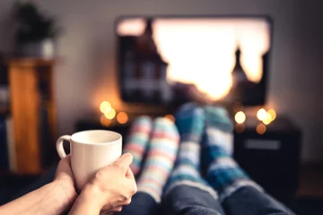 Foto op Plexiglas Couple drinking tea, hot chocolate, eggnog or mulled wine and watching tv in warm cozy woolen socks in winter. Woman holding cup of morning coffee in home living room. Sick people with flu or fever. © terovesalainen