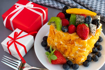Beautiful, delicious, fruit cake in white plate, red and white gift boxes, napkin, fork on grey tablecloth.