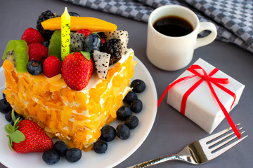 Beautiful, delicious, fruit cake with candle in white plate, white gift box, napkin, cup of coffee, fork on grey tablecloth.