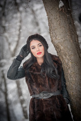 Beautiful woman in long black fur coat and cap enjoying the winter scenery in forest. Brunette girl posing under snow-covered trees branches in woods. Attractive young female in bright cold day