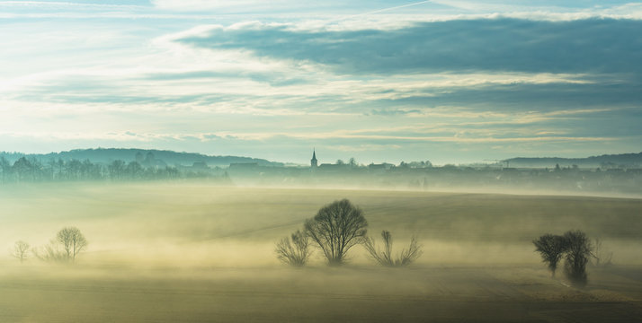 dramatic blue yellow foggy morning landscape on a winter morning in the Bavarian Utnerfranken a village in the background. fields and leafless trees as a motive. everything looks mystical
