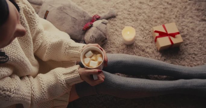 High angle shot of a woman in a knitted sweater holding a cup of hot drink with marshmallows, rocking slowly side to side near a candle and gift box. Vertical camera tilt.