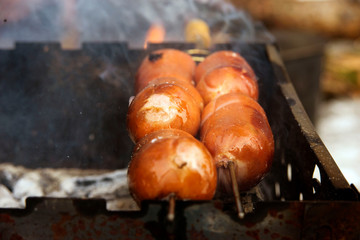 juicy, tasty and fragrant sausages pierced with a skewer with a yellow handle on the grill to roast over the coals at a picnic in the winter forest. Smoke and aroma around. Warm welcome friends.