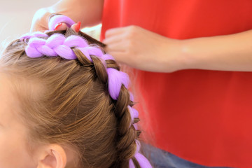 Fototapeta premium Beautiful caucasian white little girl at the professional hair salon. Hairdresser making two stylish braids with violet kanekalon for smiling adorable kid. Braided child hair with pony tails.