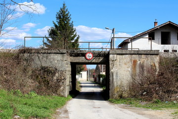 Fototapeta na wymiar Old train overpass with cracked concrete partially overgrown with dried plants without leaves with narrow paved road going through it and houses and trees with cloudy blue sky in background