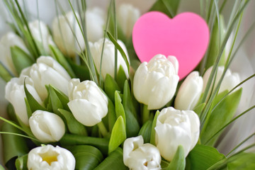 Bouquet of white beautiful tulips with tender petals and fresh green leaves with selective focus and blurred pink heart card for romantic valentines or mothers day. Spring bunch of flowers