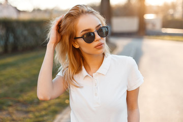 Modern stylish young hipster woman in trendy white polo t-shirt in black sunglasses stand and...