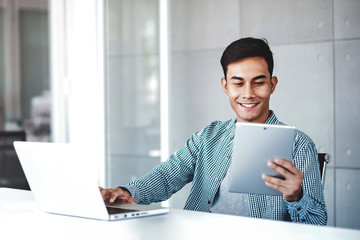 Happy Young Asian Businessman Working on Computer Laptop in his Workplace. Smiling and looking at...