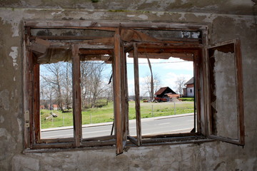 View of paved road and houses surrounded with grass and trees from inside of abandoned house through destroyed window with broken window frame mounted on cracked inner wall on warm sunny day