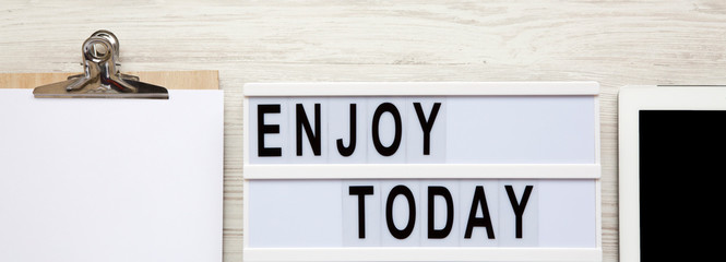 Workspace with tablet, notepad and 'Enjoy today' word on modern board over white wooden surface, top view. From above, flat-lay, overhead.