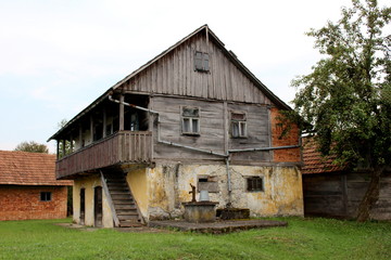 Wooden dilapidated old family house with stone foundation, broken wooden boards and large front balcony surrounded with uncut grass, well with hand water pump and metal bucket with small red brick str