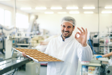 Young Caucasian worker in work wear holding tray with cookies and shoving okay sign while standing...