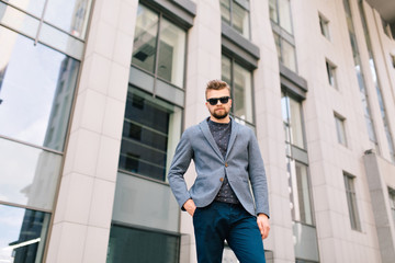 Handsome guy with beard is standing outdoor  on office building background. He wears gray jacket, jeans, sunglasses. He looks to the camera. Bottom view.