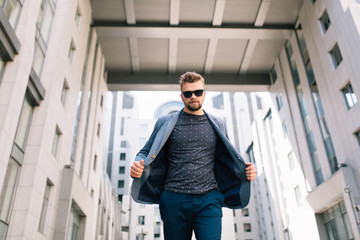 Attractive man with beard is walking to the camera outdoor on office building background. He wears T-shirt, jeans, sunglasses. He takes off his jacket. Bottom view.