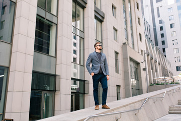 Handsome guy in sunglasses is standing on office building background. He wears gray jacket, jeans, brown shoes. He keeps hands in pockets.