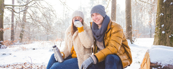 Glückliches Paar im winterlichen Wald sitzt auf einem Baumstamm und trinkt Tee