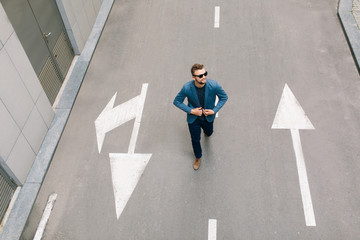 Handsome man in sunglasses is walking on street. He wears gray jacket, jeans, brown shoes, beard....
