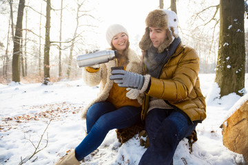 Obraz premium Glückliches Paar im winterlichen Wald sitzt auf einem Baumstamm und trinkt Tee