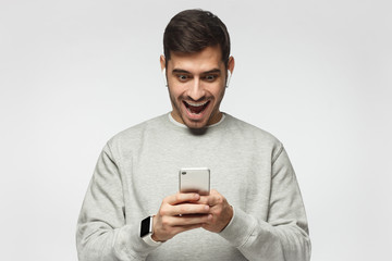 Young excited man looking at his phone with surprise expression, isolated on gray background