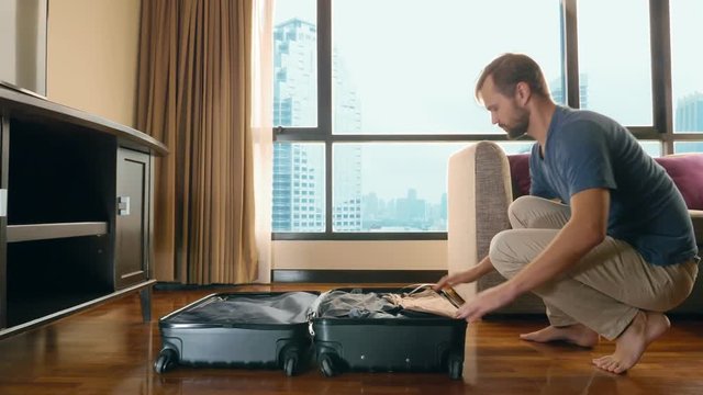 handsome man packs a suitcase in a room with a panoramic window overlooking the skyscrapers
