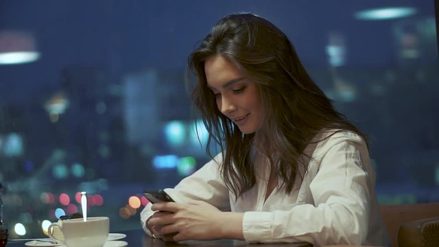 young girl is texting a message on the smartphone while sitting in a cafe on the background of the lights of the night city