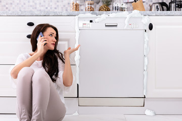 Sad Woman Sitting Next To Damaged Dishwasher Calling Technician