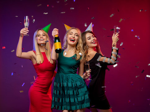 Portrait of three amazing, cute girls in fashion dresses and party caps, holding glass with champagne, celebrating the New year or Birthday party, standing over colorful background