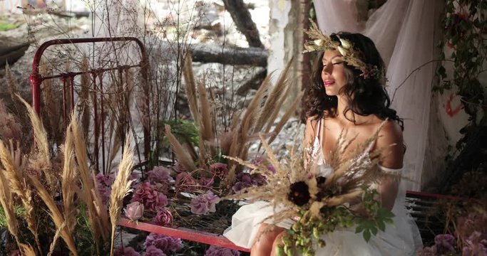 Conceptual shooting. 4k. Woman with long dark hair dressed like a nymph sits on the old bed among blooming pink flowers and hau and smiles looking straight in the camera