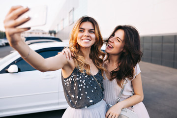 Portrait two pretty girls having fun on parking. Blonde girl making selfie-portrait, brunette near showing tongue, they smiling to camera