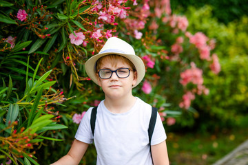 Blonde boy in the straw hat and big glasses staying among green trees and bushes with pink flowers