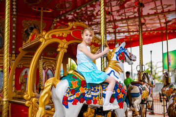 Fototapeta na wymiar Blonde girl with two braids in white and blue dress riding colorful horse in the merry-go-round carousel in the entertainment park