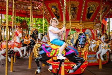 Fototapeta na wymiar Blonde boy in the straw hat and big glasses riding colorful horse in the merry-go-round carousel in the entertainment park