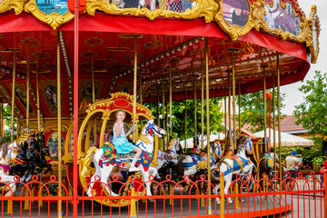 Blonde girl with two braids in white and blue dress riding colorful horse in the merry-go-round carousel in the entertainment park